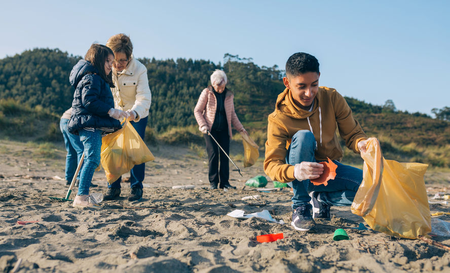 four people picking up trash in the sand on a beach, one in front is bent and has a plastic bottle in his hand