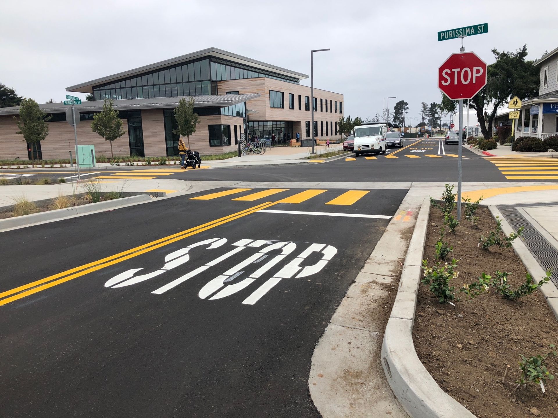 fresh painted street with a crosswalk and the word "Stop" painted on the street; to the left is a large building with windows, to the right there's dirt and shrubs in between a sidewalk and the street